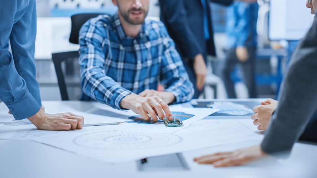 Engineers and business executives gathered around a PCB and drawings discussing design for supply chain topics