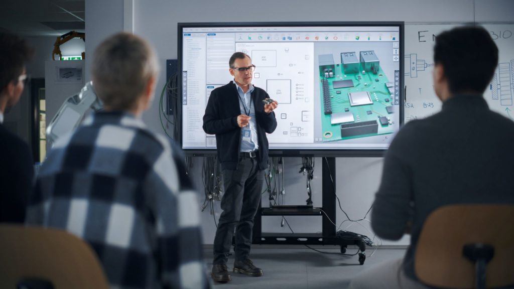 Diverse team of engineers watching a meeting room monitor displaying a printed circuit board model.